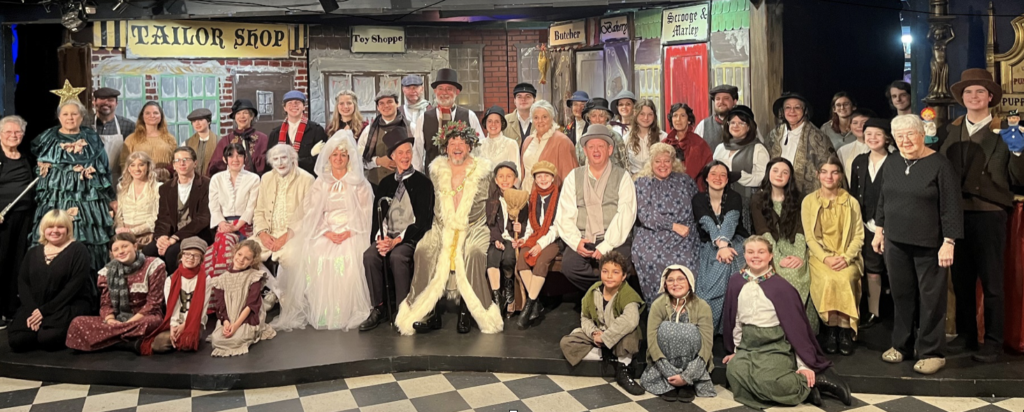 50 people in holiday costumes pose on a stage decorated for the musical "Scrooge." 